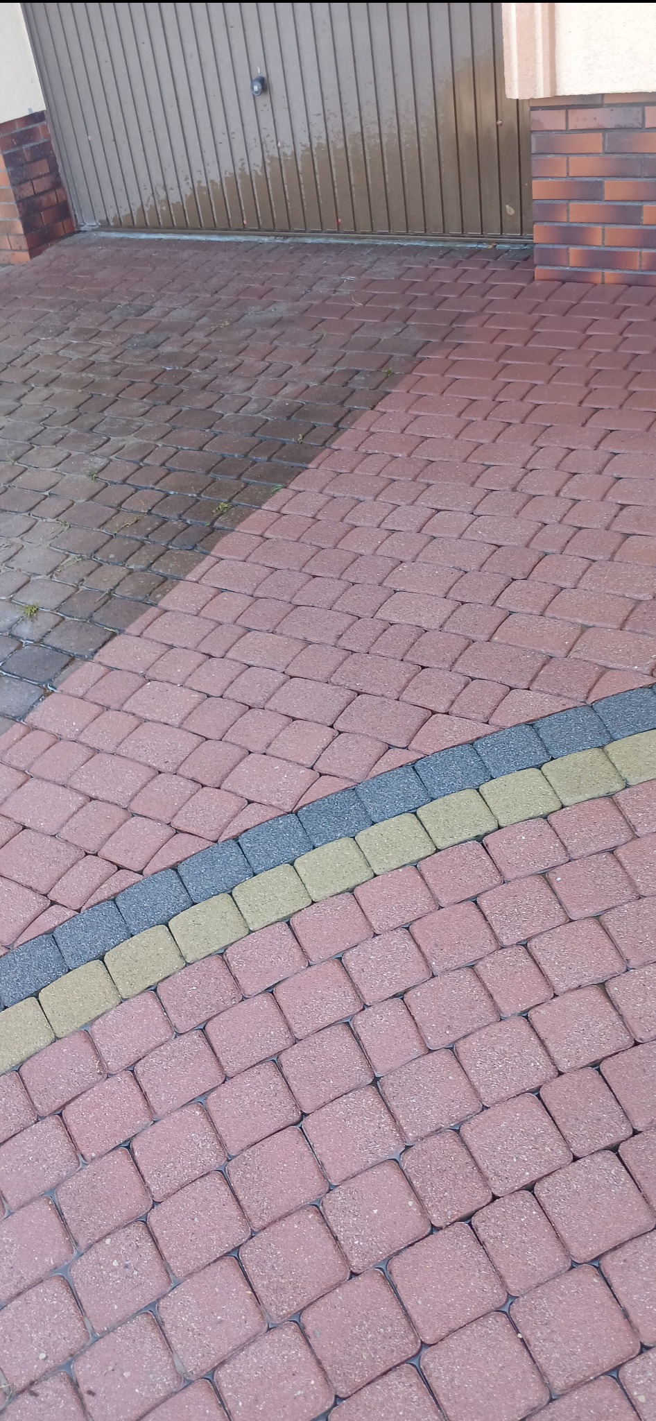 Częściowo umyta, czerwona kostka brukowa przed garażem, z widoczną różnicą w czystości, z dekoracyjnym pasem z szarej i żółtej kostki.