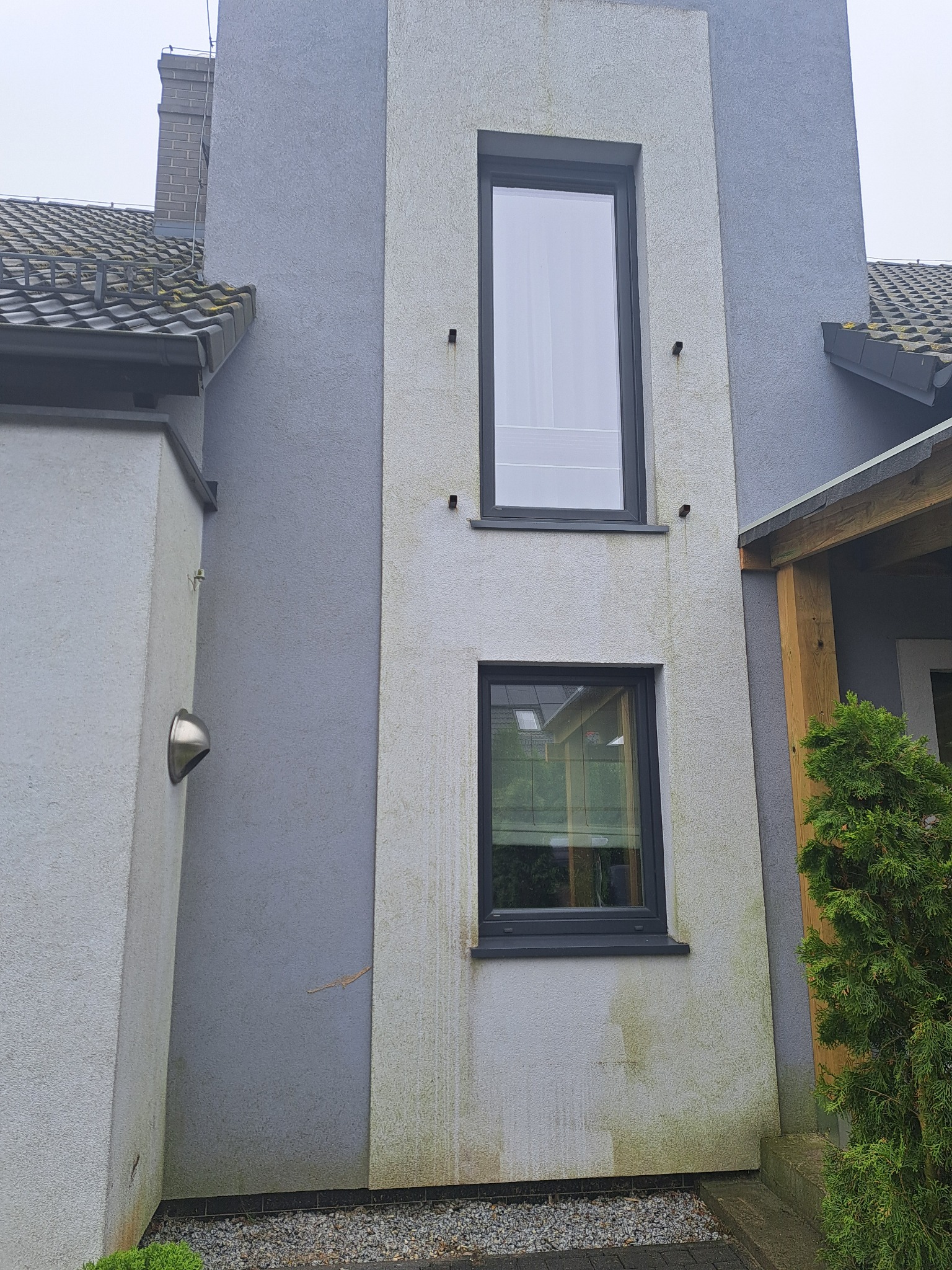 Szara elewacja dwukondygnacyjnego domu z dwoma oknami, widoczne zabrudzenia na jasnym fragmencie ściany i zielony mech na dachu.