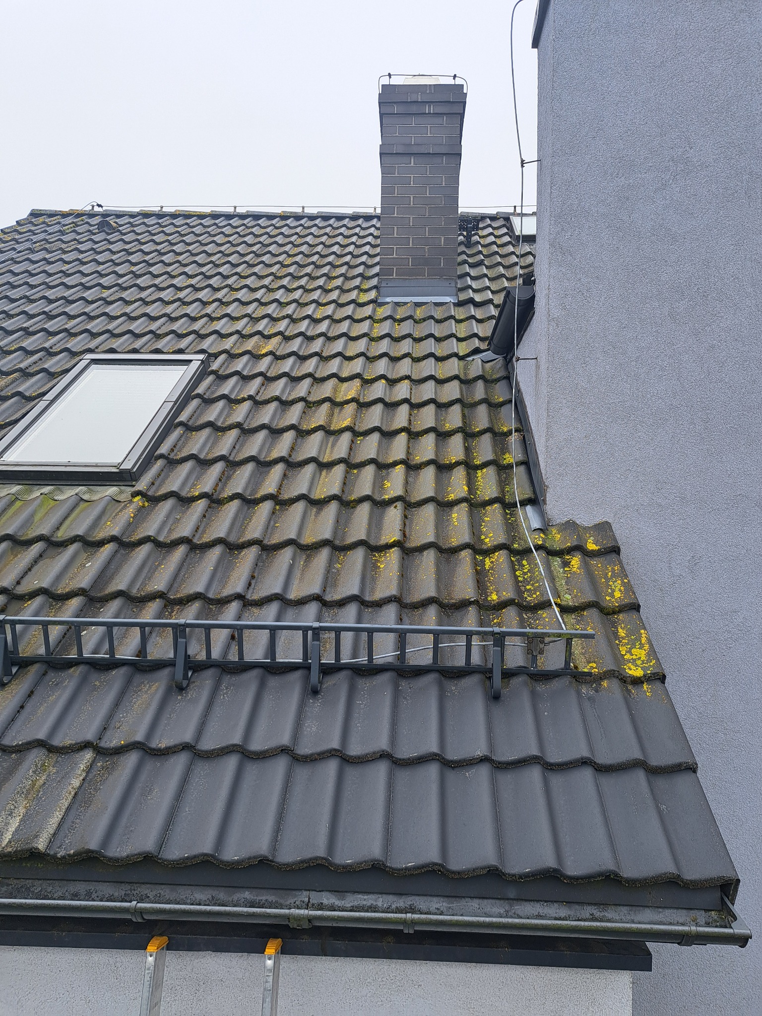 Ciemna dachówka z mchem, widoczny komin i okno dachowe. Fragment drabiny w dolnej części kadru. Ujęcie z dołu, pochmurne niebo w tle.