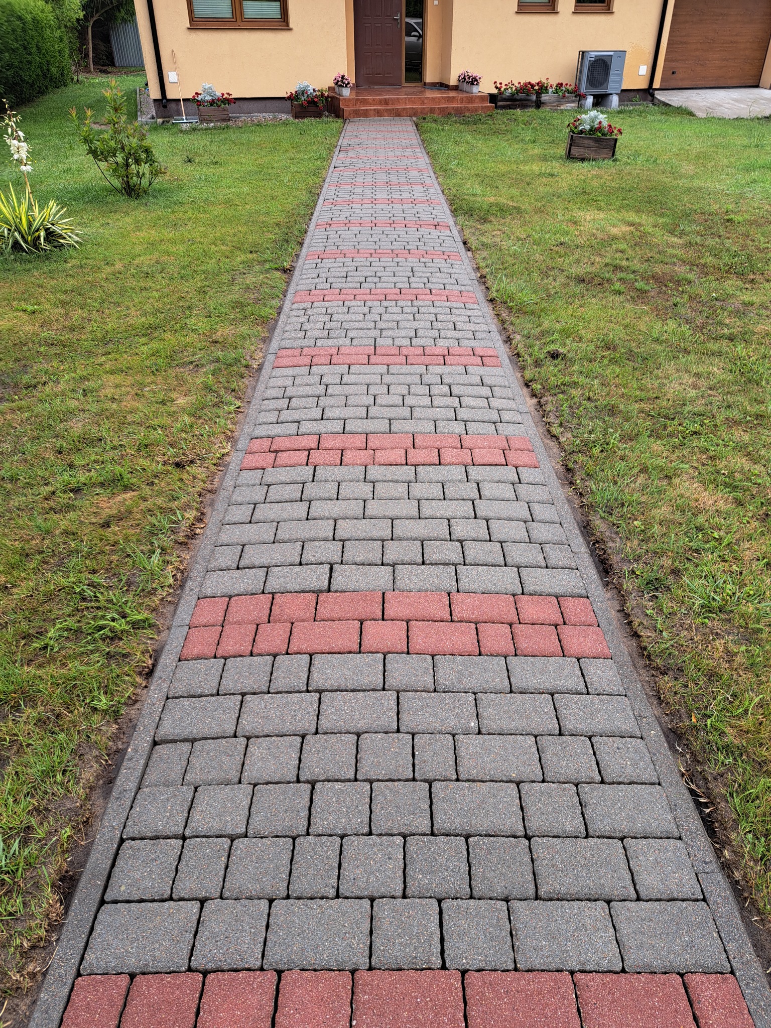 Ścieżka z kostki brukowej w odcieniach szarości i czerwieni prowadząca do domu z zielonym trawnikiem po bokach, rabatki z kwiatami.