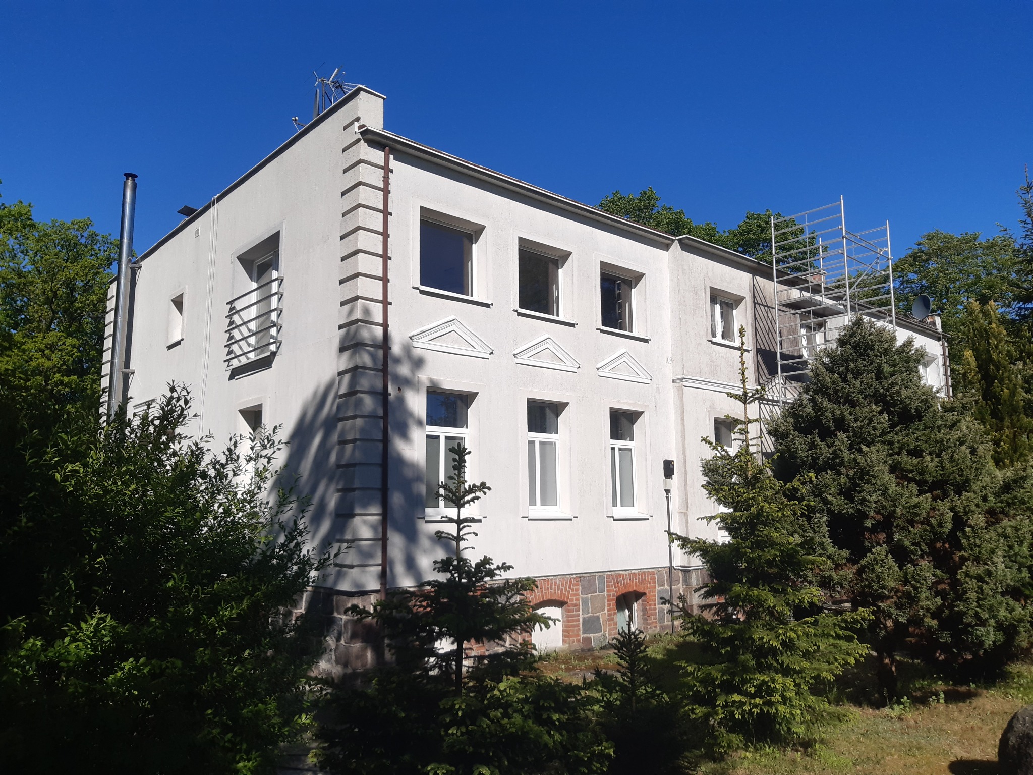 Dwupiętrowy, jasny budynek z elementami dekoracyjnymi, otoczony zielenią. Widoczne rusztowania przy ścianie, sugerujące prace remontowe lub czyszczenie elewacji.