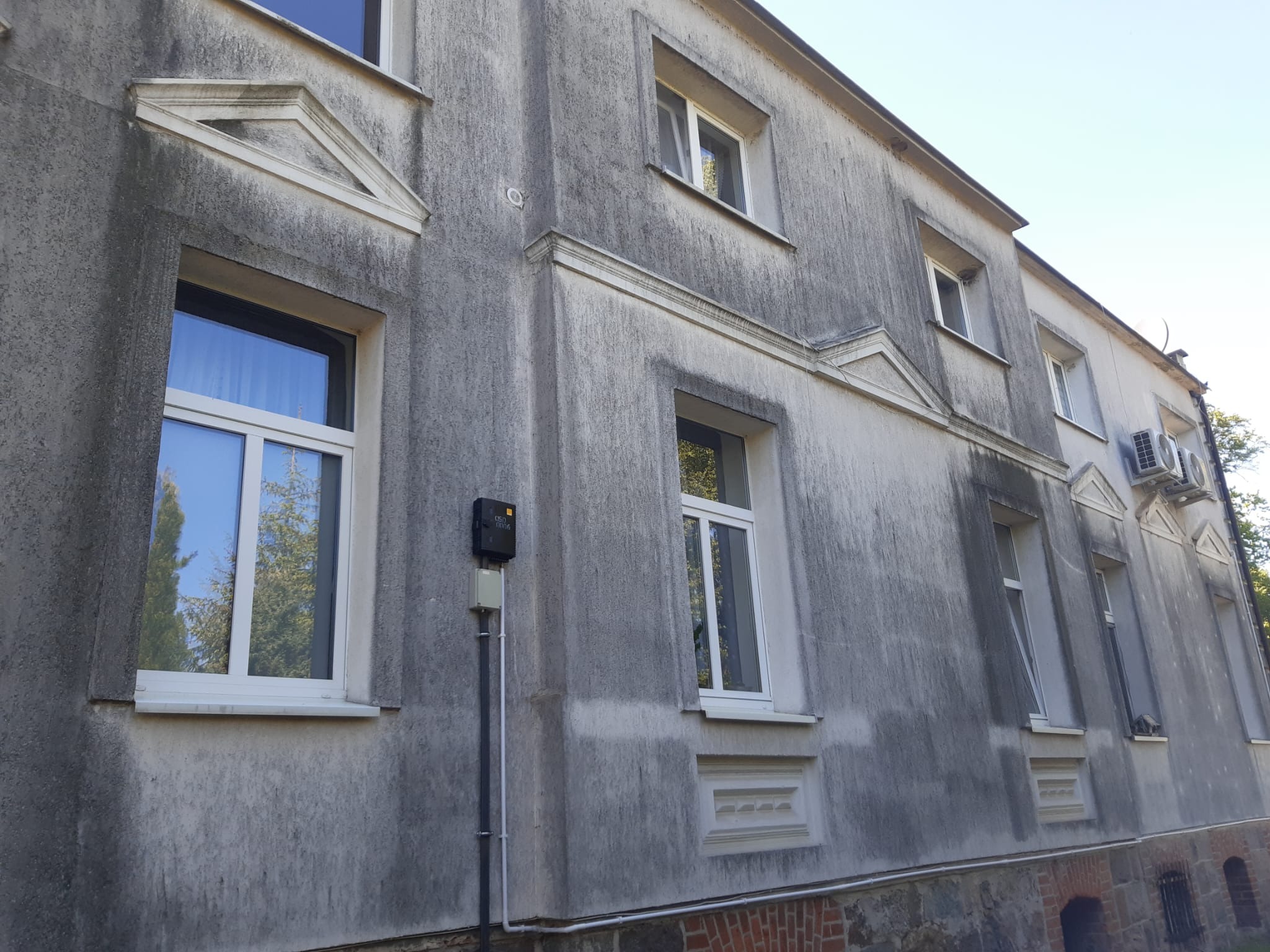 Szara elewacja budynku z widocznymi zabrudzeniami i zaciekami. Widoczne okna z białymi ramami, elementy dekoracyjne i instalacje na ścianie.
