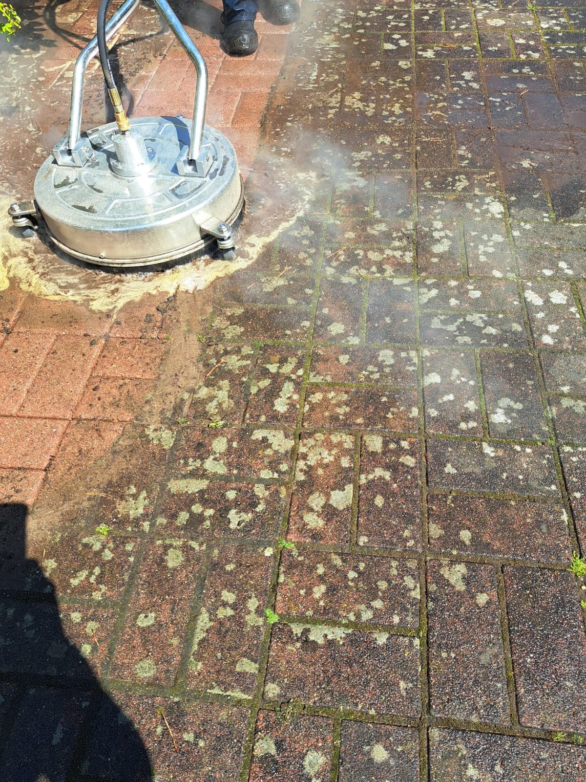 Ceglana kostka brukowa czyszczona myjką ciśnieniową, widoczna różnica między umytą a zabrudzoną powierzchnią. W tle osoba w czarnych butach.