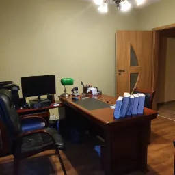 Biuro z drewnianym biurkiem, lampką bankierską, komputerem i rzędem niebieskich segregatorów, oświetlone ozdobnym żyrandolem.