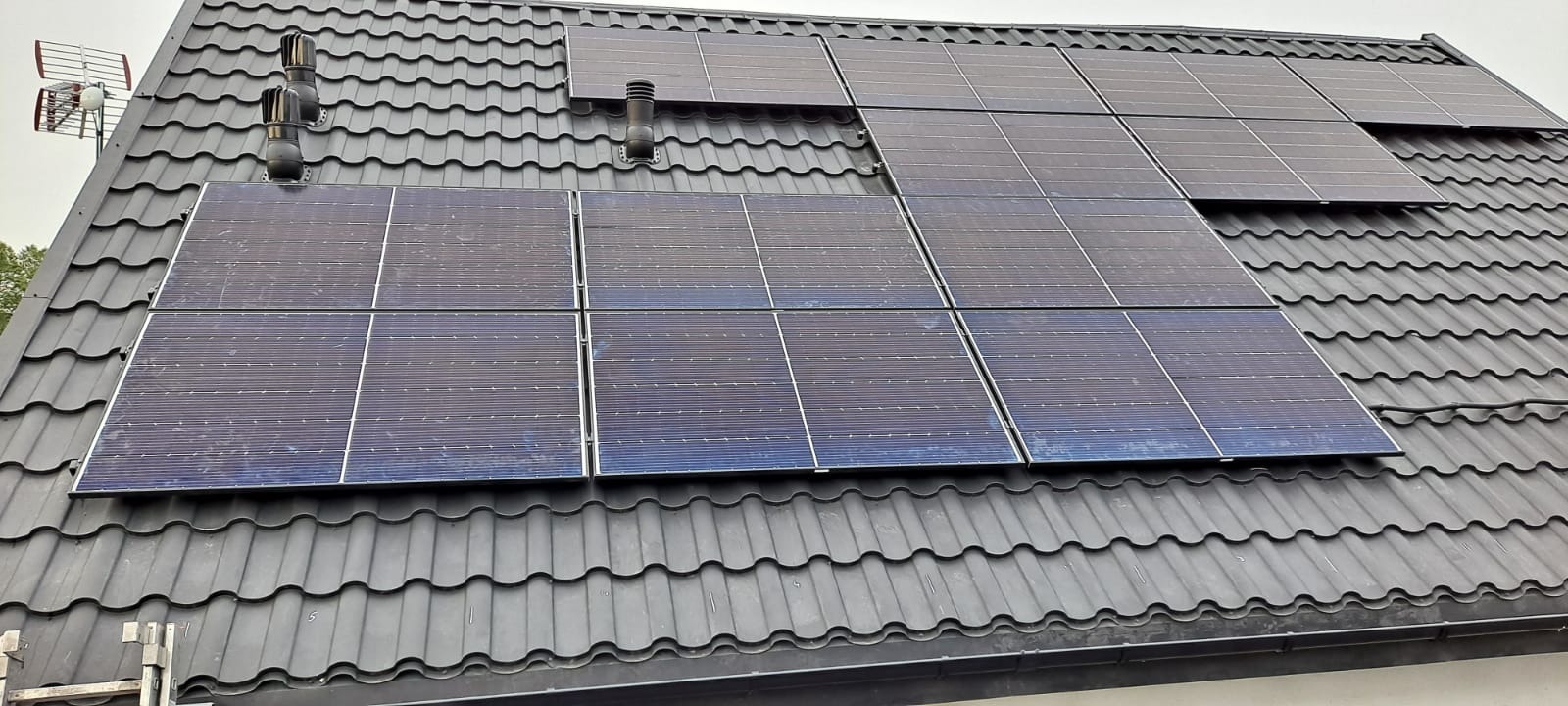 Częściowo zamontowane panele słoneczne na ciemnoszarym dachu z dachówki ceramicznej, widoczny komin wentylacyjny i antena telewizyjna.