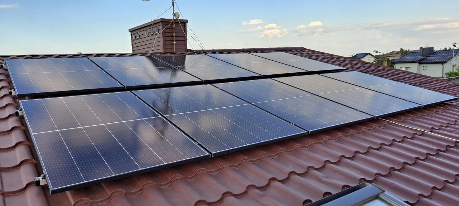 Panele słoneczne zamontowane na dachu z brązowej dachówki ceramicznej, widoczny komin w tle i błękitne niebo.