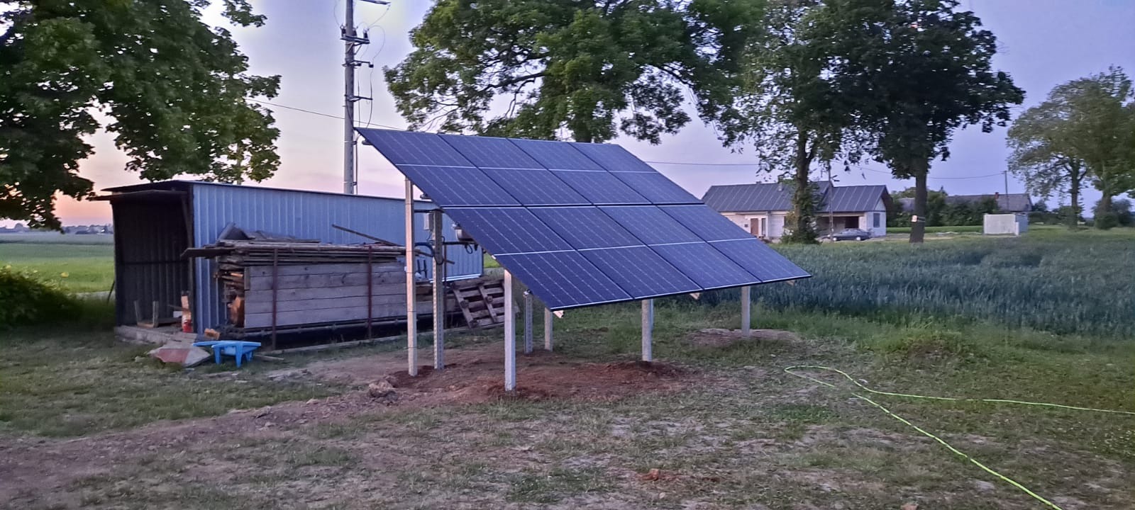 Panele słoneczne zamontowane na metalowych słupkach na tle wiejskiego krajobrazu z budynkami i polami.