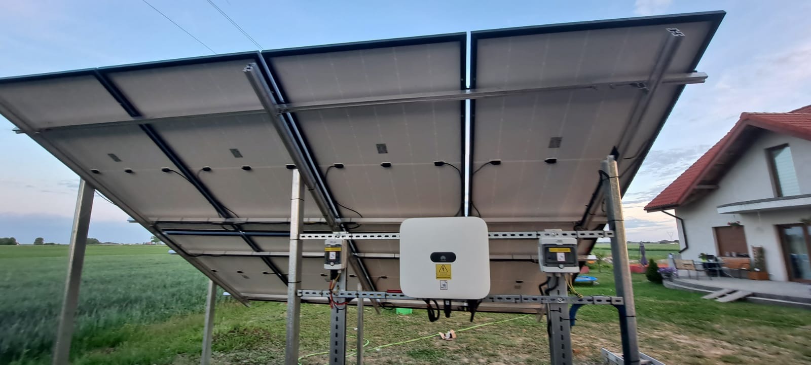Widok od spodu na panele słoneczne zamontowane na metalowej konstrukcji, z widocznym inwerterem Huawei i elementami instalacji elektrycznej, na tle zielonego pola i domu jednorodzinnego.
