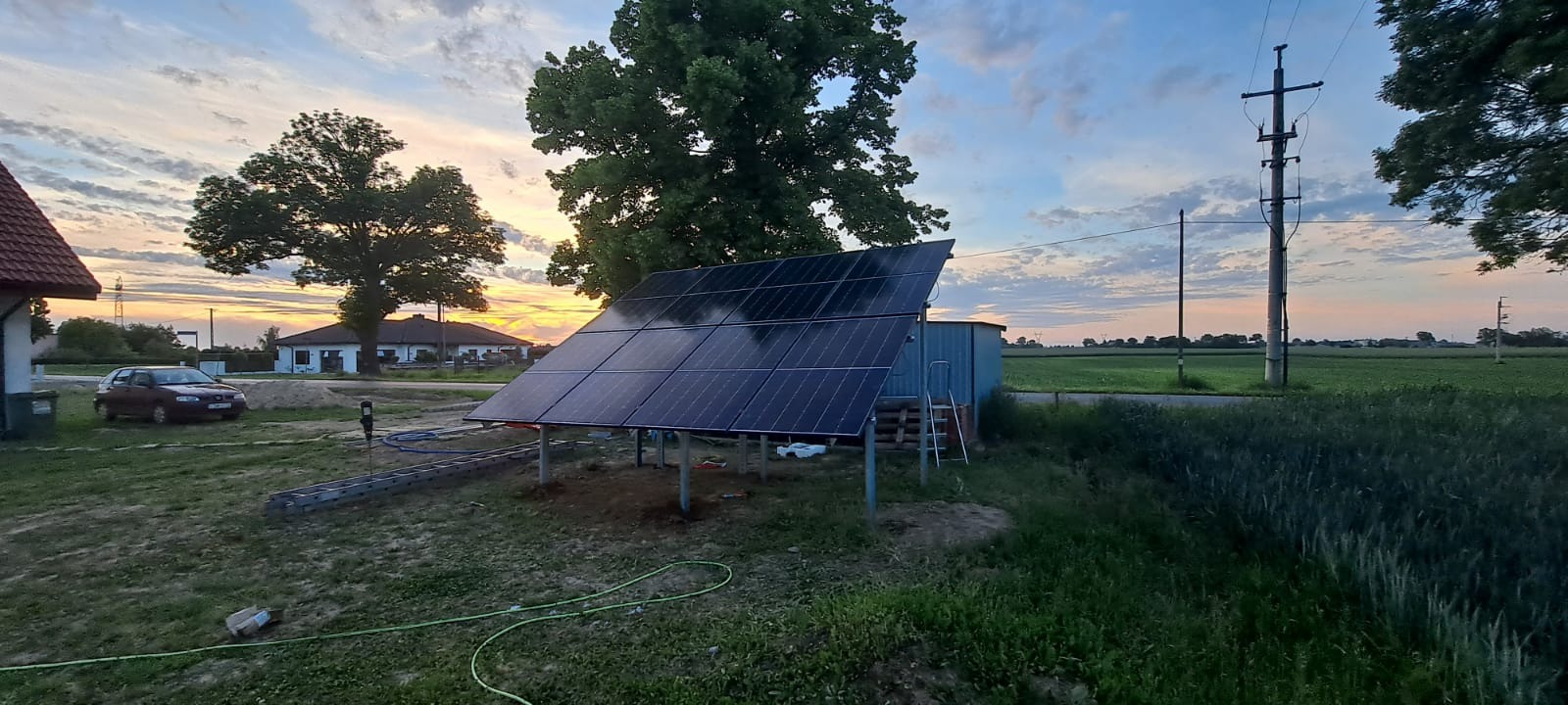 Panele fotowoltaiczne na metalowej konstrukcji w ogrodzie, widoczne przewody i drabina, w tle dom i zachodzące słońce.