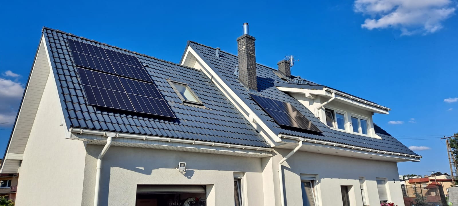 Panele słoneczne zamontowane na dachu domu jednorodzinnego z szarej dachówki, widoczne kominy i okno dachowe, błękitne niebo z chmurami w tle.