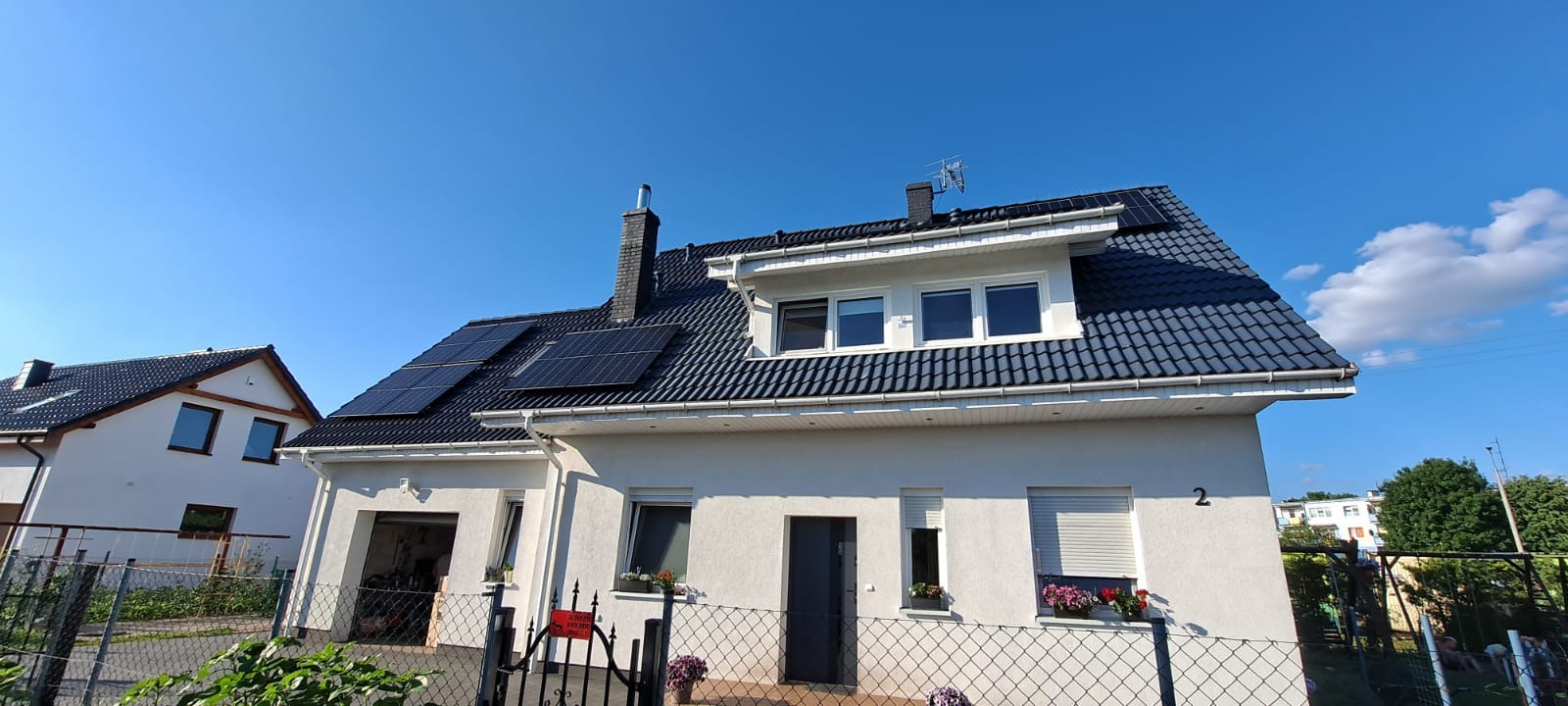 Biały dom jednorodzinny z ciemną dachówką, na której zamontowane są panele fotowoltaiczne; w tle błękitne niebo i fragment drugiego domu.