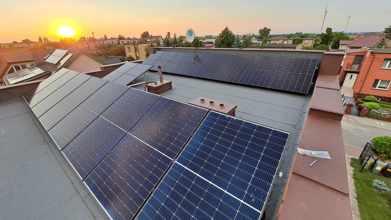 Instalacja paneli słonecznych na płaskim dachu budynku mieszkalnego, widok z góry, narzędzia montażowe leżące obok paneli, zachodzące słońce w tle.