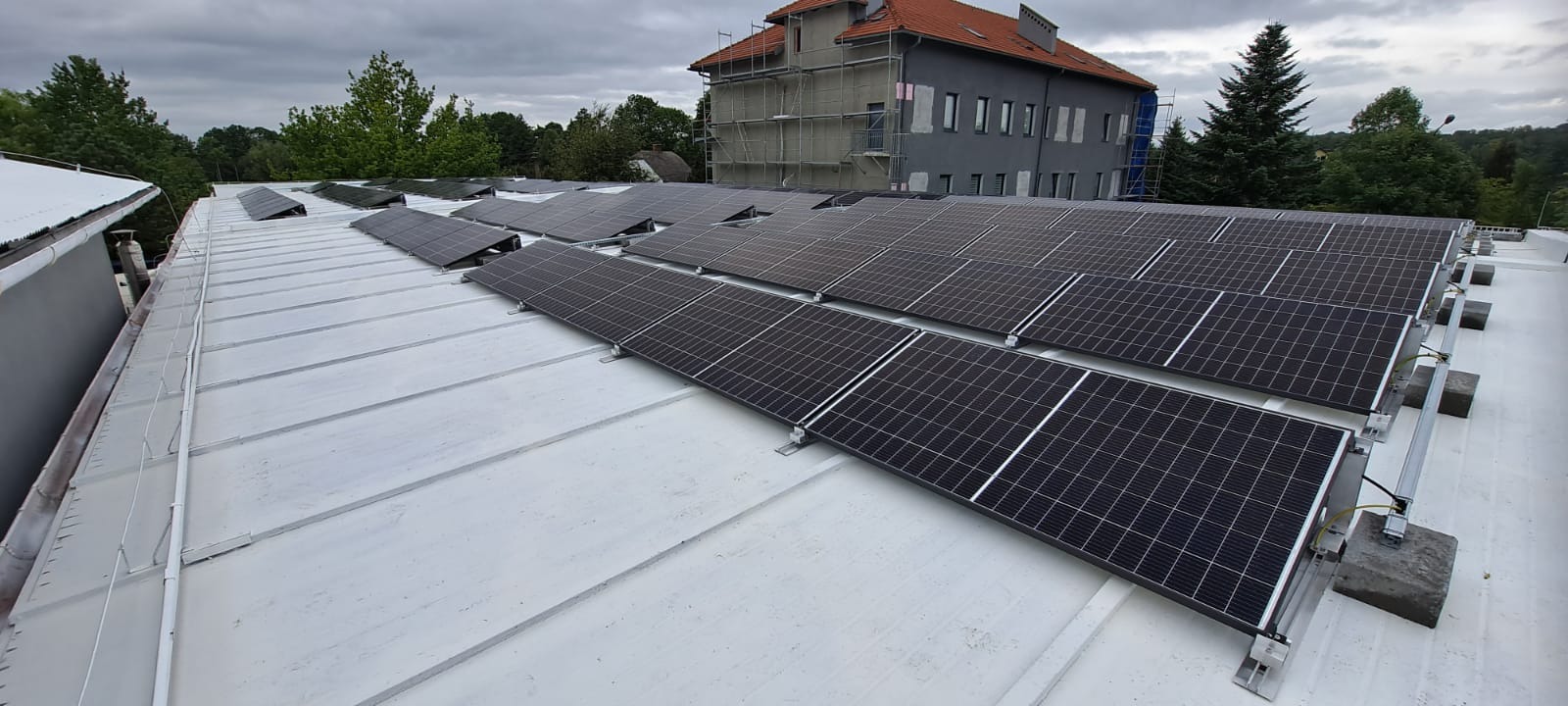 Rząd paneli słonecznych na płaskim, białym dachu budynku, z widocznym budynkiem w tle w trakcie remontu elewacji.