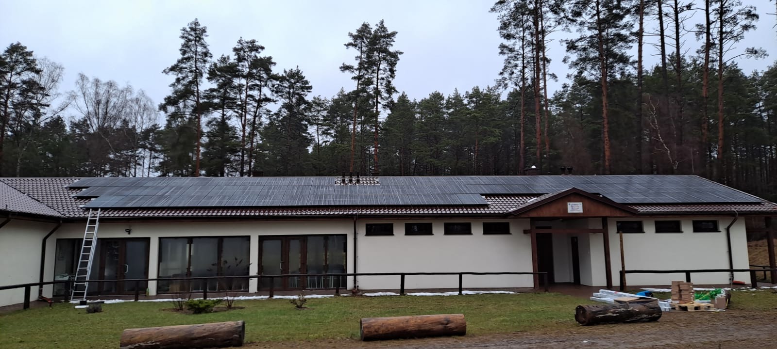 Instalacja fotowoltaiczna na dachu budynku z białymi ścianami, widoczne panele słoneczne w czarnych ramach, tło stanowią drzewa, na ziemi leżą drewniane kłody i materiały budowlane, po lewej...