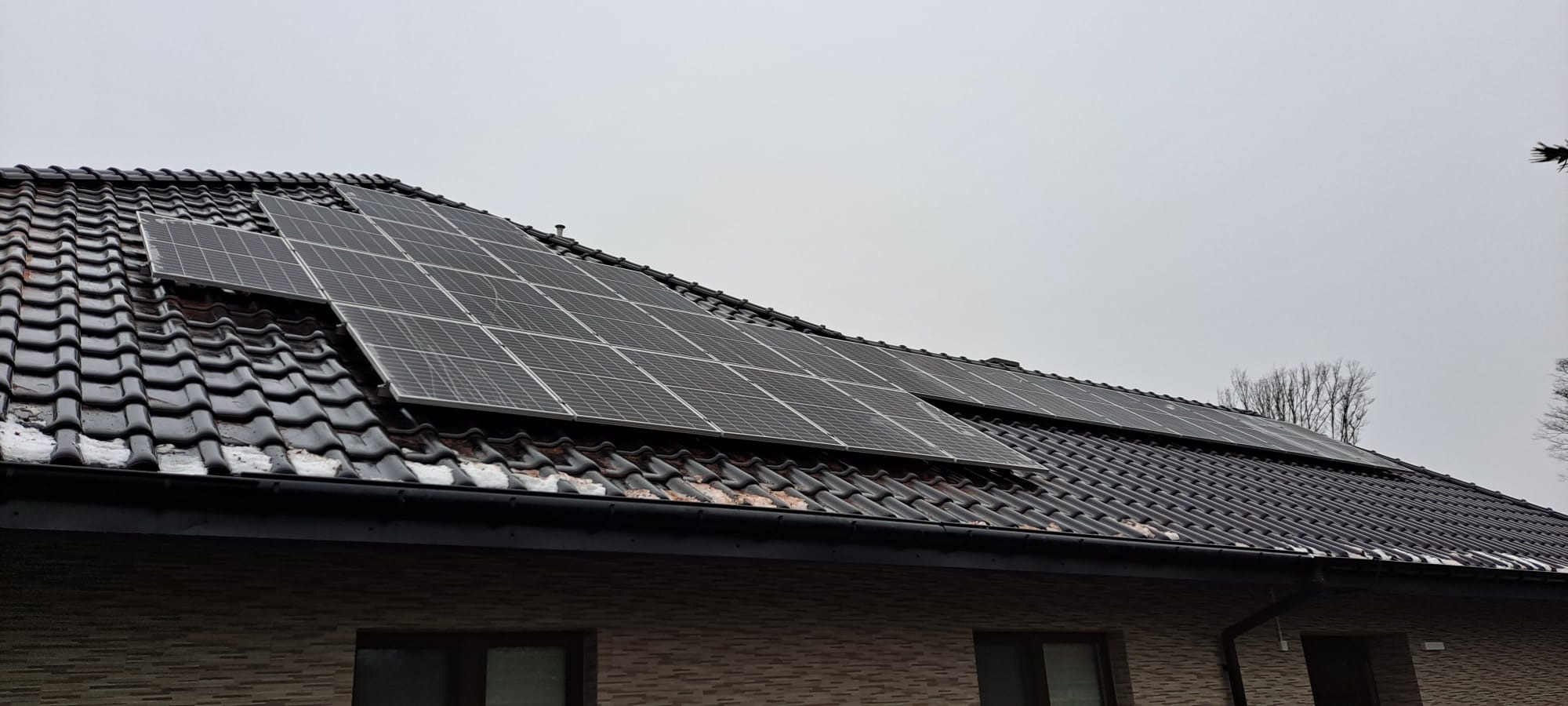 Panele słoneczne na ciemnym, ceramicznym dachu domu jednorodzinnego w pochmurny dzień, widoczne ślady topniejącego śniegu na dachówkach.