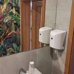 Nowoczesna łazienka z umywalką, armaturą, dozownikiem mydła w płynie i lustrem, w którym odbija się fototapeta z motywem roślinnym i dozownik ręczników papierowych.