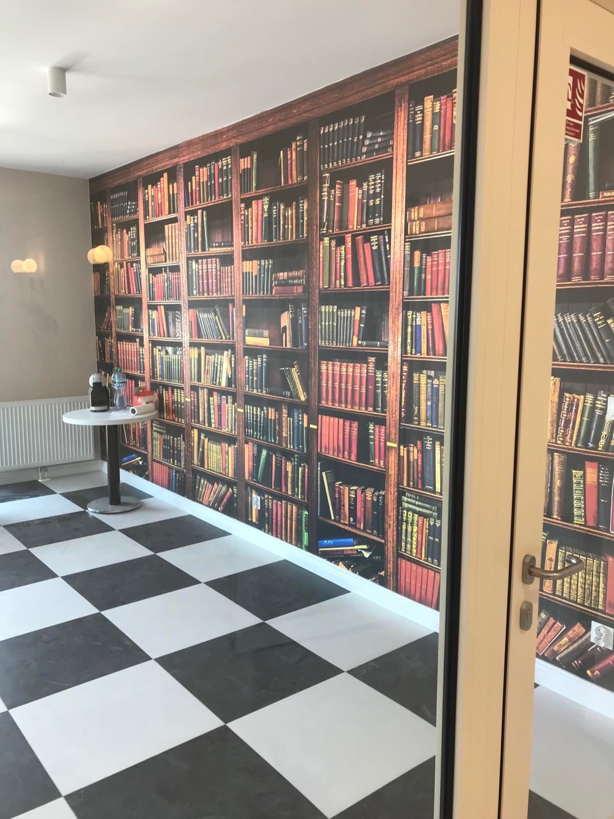 Pokój z tapetą imitującą biblioteczkę, czarno-biała szachownica na podłodze i okno z widoczną częścią tapety.