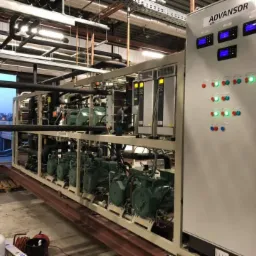 Rozbudowana instalacja chłodnicza Advansor z wieloma sprężarkami i panelem sterowania z wyświetlaczami, zainstalowana w pomieszczeniu technicznym, widoczne rurociągi i elementy konstrukcyjne budynku.