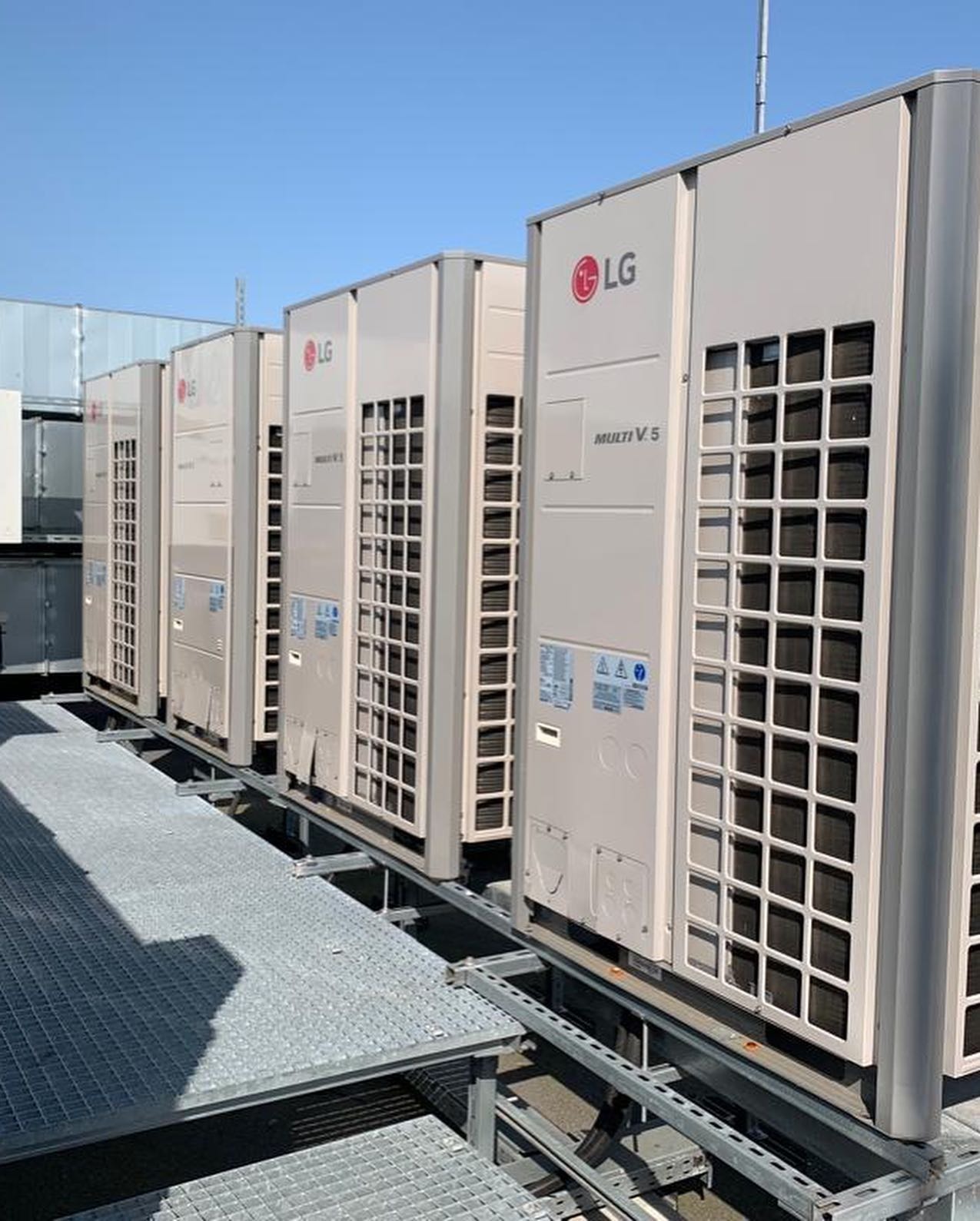 Rząd zewnętrznych jednostek klimatyzacyjnych LG Multi V 5 ustawionych na metalowej platformie na dachu, błękitne niebo w tle.