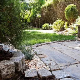 Ogród z drewnopodobnym tarasem obramowanym kamieniem i żwirem, z zielonym trawnikiem i krzewami w tle, w słoneczny dzień.