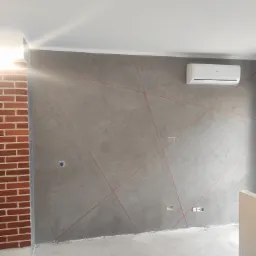Szpachlowana ściana z dekoracyjnymi, geometrycznymi liniami w kolorze czerwonym, obok fragmentu ściany z cegły, z widocznym klimatyzatorem i gniazdkami elektrycznymi, w trakcie prac wykończeniowych.