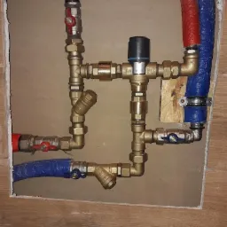 Kompleksowy węzeł instalacyjny z mosiężnymi kształtkami, zaworami kulowymi z czerwonymi i niebieskimi motylkami, filtrem skośnym i rurami w izolacji, zamontowany w otworze w ścianie wykończonej...