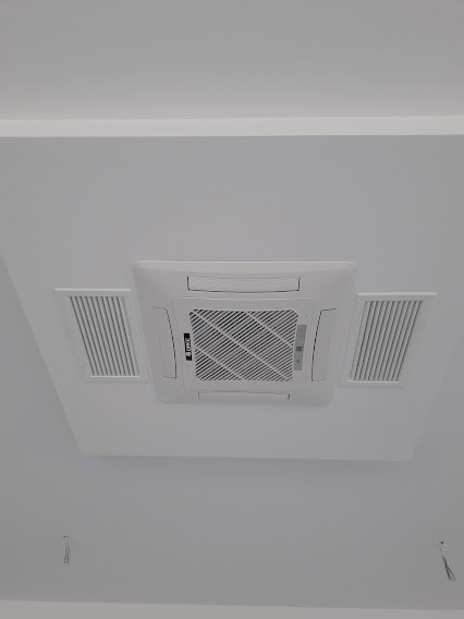 Kasetonowa jednostka klimatyzacyjna zamontowana w podwieszanym suficie, widoczne kratki wentylacyjne po bokach. Białe wykończenie i minimalistyczny design.