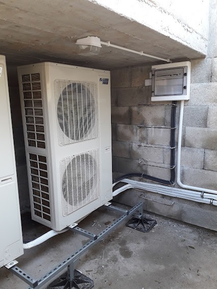 Dwie jednostki zewnętrzne klimatyzacji na konstrukcji wsporczej, widoczna instalacja elektryczna i oświetlenie, ściana z betonowych bloczków.