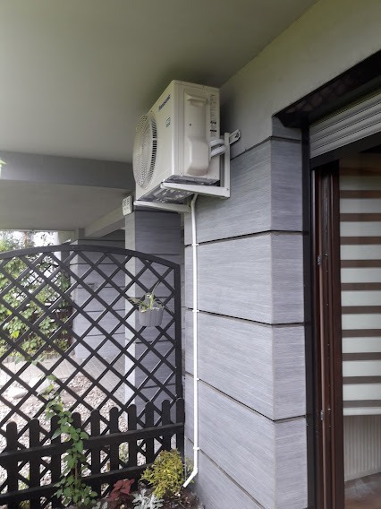 Zewnętrzna jednostka klimatyzacji Panasonic zamontowana na ścianie budynku z szarą elewacją, widoczna biała rurka odprowadzająca skropliny, obok drewniana kratka z roślinami.