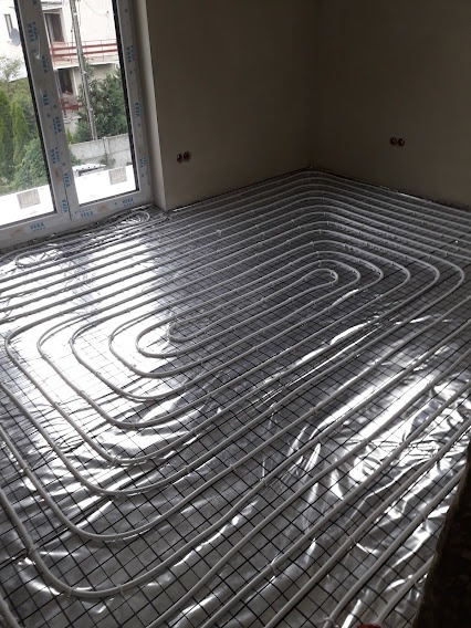 Białe rurki ogrzewania podłogowego ułożone spiralnie na izolacji z folii aluminiowej i siatce w nowym budownictwie, widok z góry na fragment podłogi w jasnym pomieszczeniu z dużym oknem.