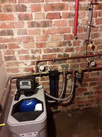 Zamontowany zmiękczacz wody Water Technic z niebieskim zbiornikiem, widoczne miedziane rury z manometrami i filtrem, na tle ściany z czerwonej cegły z widocznymi spoinami.