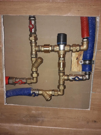 Kompleksowy węzeł instalacyjny z mosiężnymi kształtkami, zaworami kulowymi z czerwonymi i niebieskimi motylkami, filtrem skośnym i rurami w izolacji, zamontowany w otworze w ścianie wykończonej...