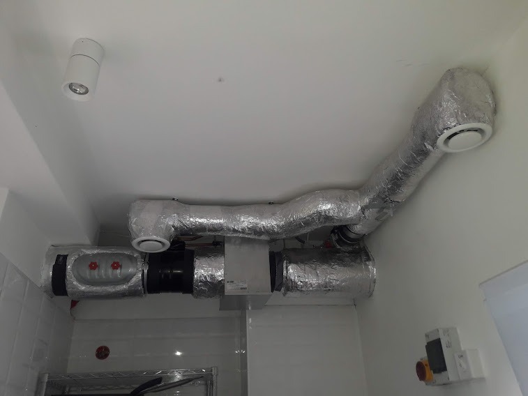 System kanałów wentylacyjnych owiniętych folią aluminiową, zamontowany pod sufitem w pomieszczeniu z białymi płytkami i oświetleniem punktowym.