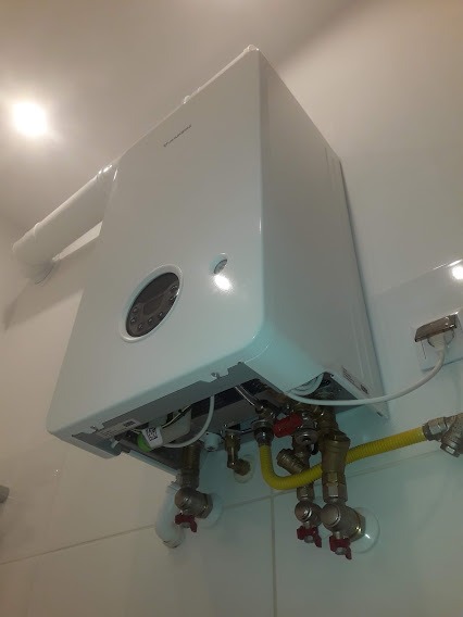 Biały, wiszący piec z widocznymi przyłączami gazowymi i wodnymi, zamontowany na tle białych płytek. Żółty przewód gazowy łączy się z mosiężnymi zaworami.