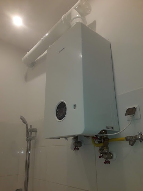 Biały, wiszący piec gazowy marki Immergas z podłączonymi rurami gazowymi i odprowadzaniem spalin, zainstalowany w łazience z widocznym prysznicem.