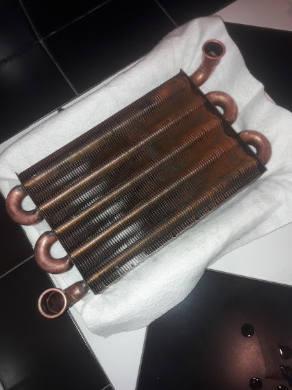 Miedziany wymiennik ciepła z widocznymi rurkami przyłączeniowymi, leżący na białym ręczniku na tle czarno-białej podłogi.