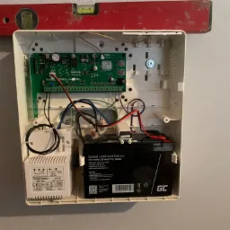 Otwarta obudowa centrali alarmowej z widoczną elektroniką, akumulatorem i transformatorem, poziomica na górnej krawędzi obudowy.