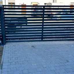 Nowoczesna, pozioma brama wjazdowa z automatyką, szara kostka brukowa na podjeździe, widok na budynek mieszkalny i zaparkowane pojazdy w tle.