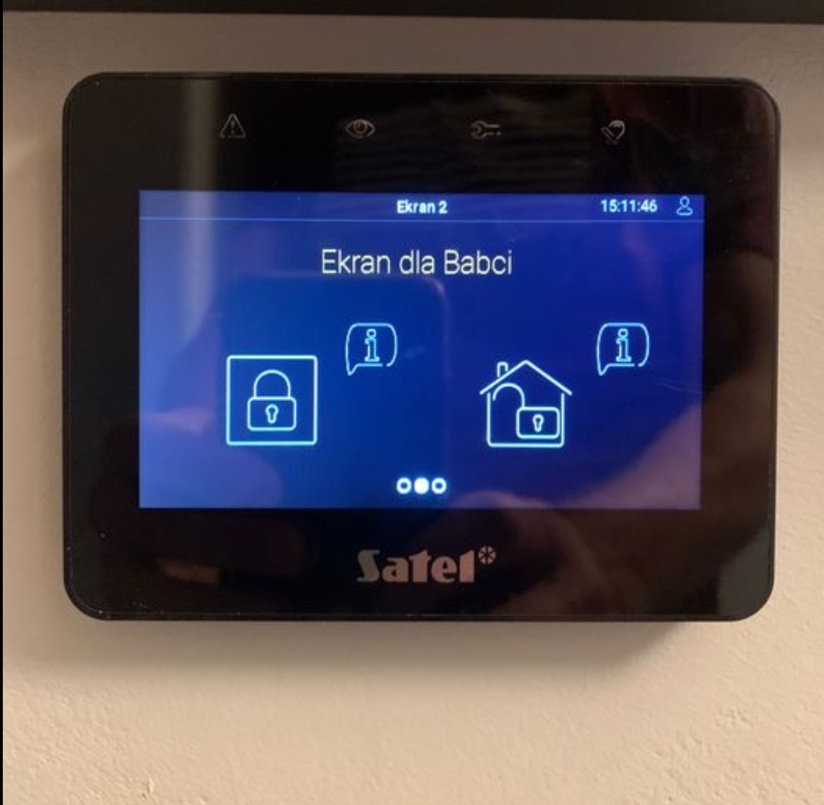 Czarny panel dotykowy Satel z interfejsem 'Ekran dla Babci', wyświetlający ikony kłódki i domu z kłódką na niebieskim tle.