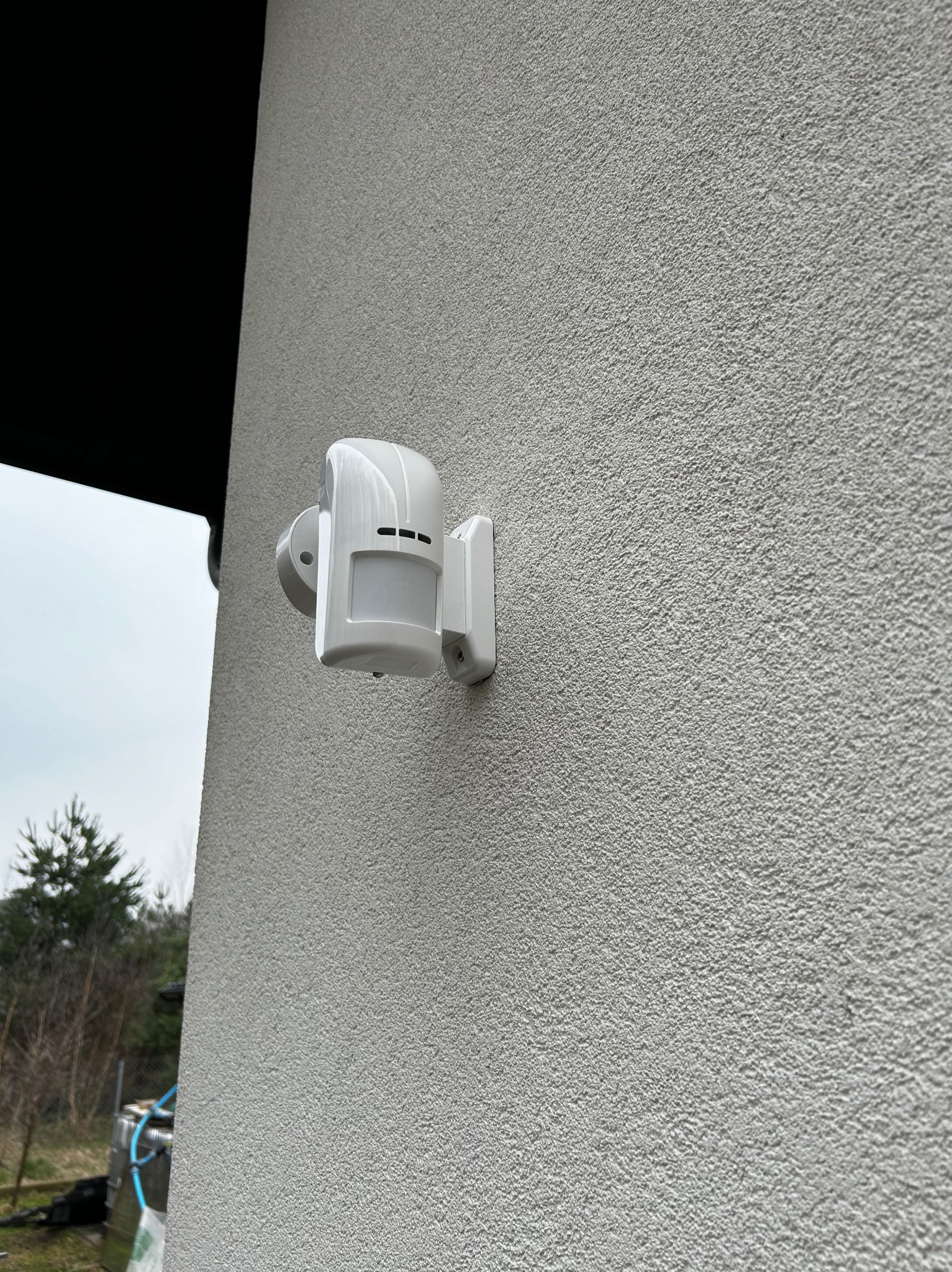 Biały czujnik ruchu zamontowany na elewacji budynku o szarej, chropowatej strukturze, widok z dołu.