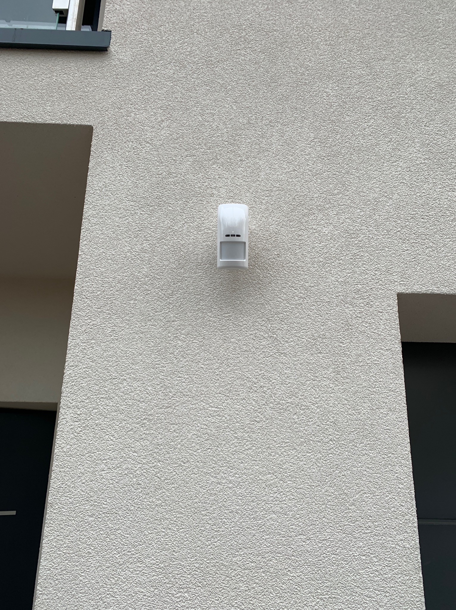 Zewnętrzny czujnik ruchu, zamontowany na elewacji budynku o jasnej, ziarnistej strukturze, widoczny fragment okna z ciemną ramą.