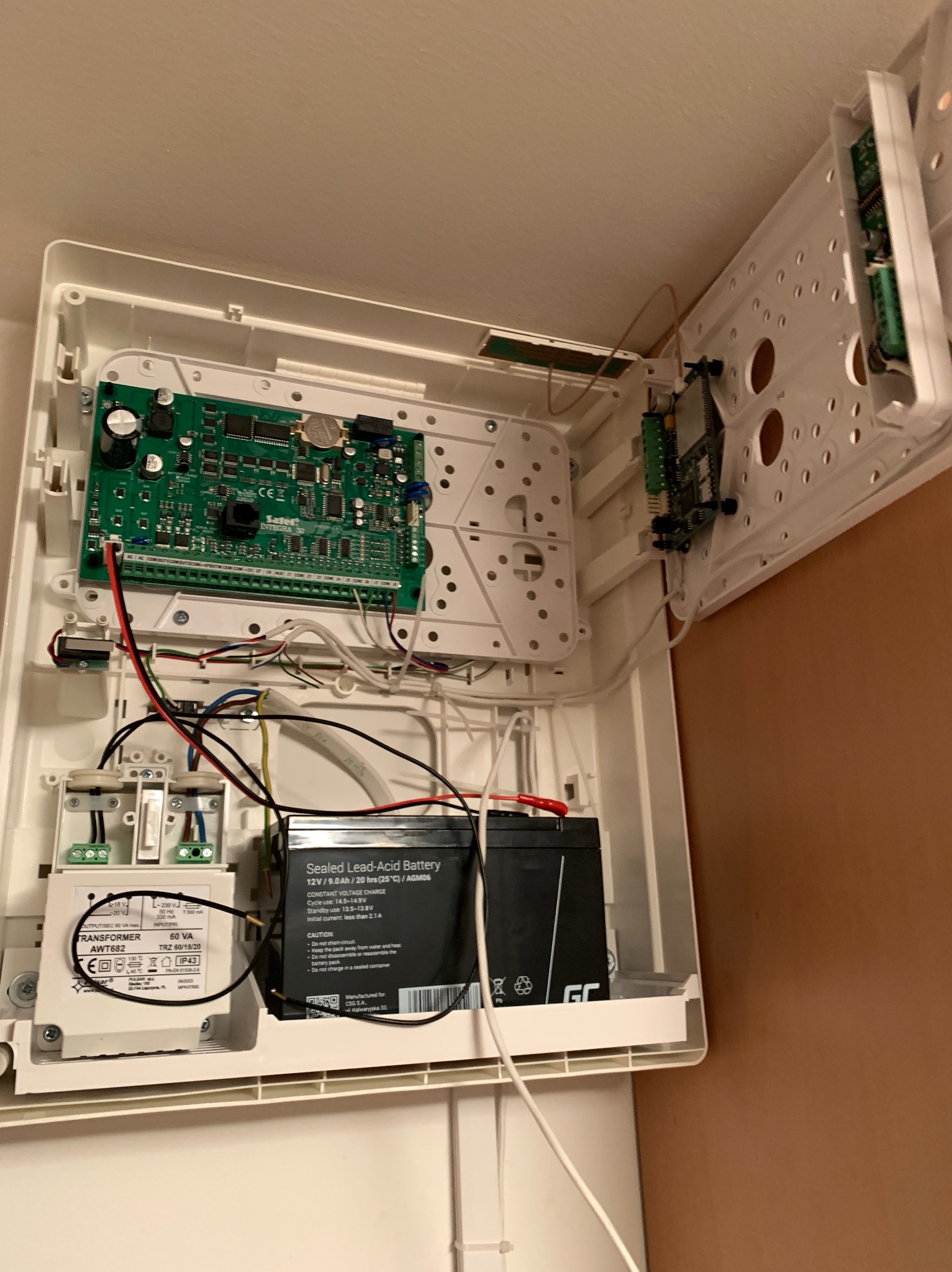 Otwarta obudowa centrali alarmowej z widoczną płytą główną, transformatorem AWT682, akumulatorem żelowym 12V/9.0Ah oraz okablowaniem.