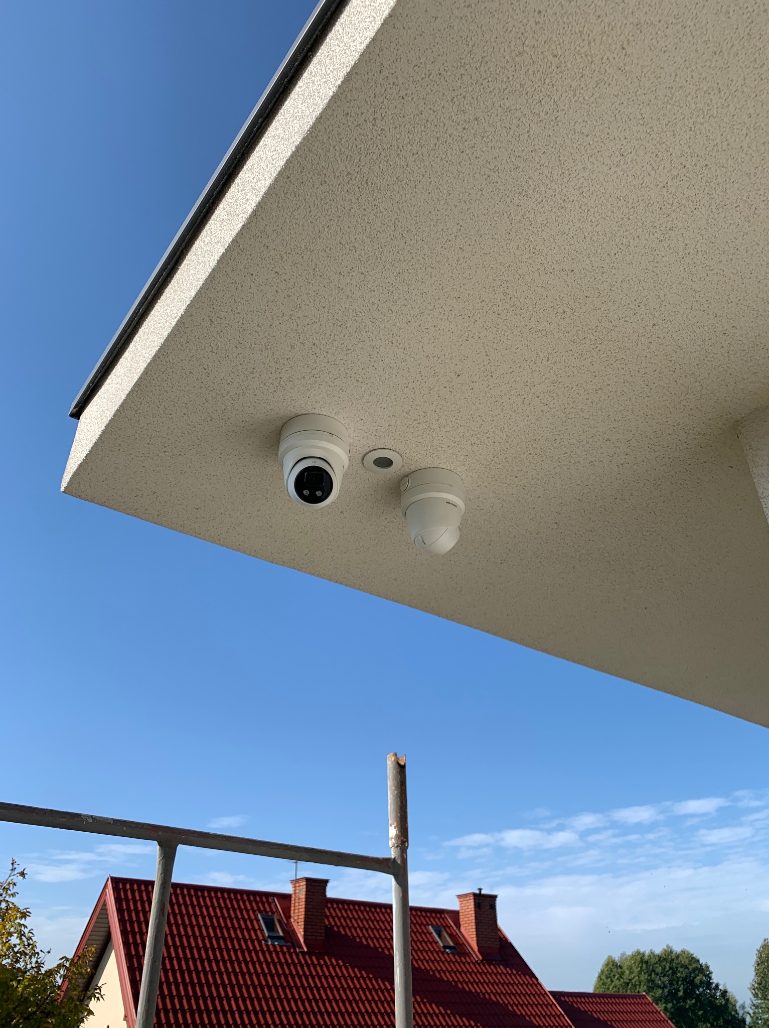 Dwie kamery monitoringu, kopułkowa i obrotowa, zamontowane pod kremowym podbitkiem w Mrozach, z widokiem na błękitne niebo i dach domu z czerwonej dachówki.