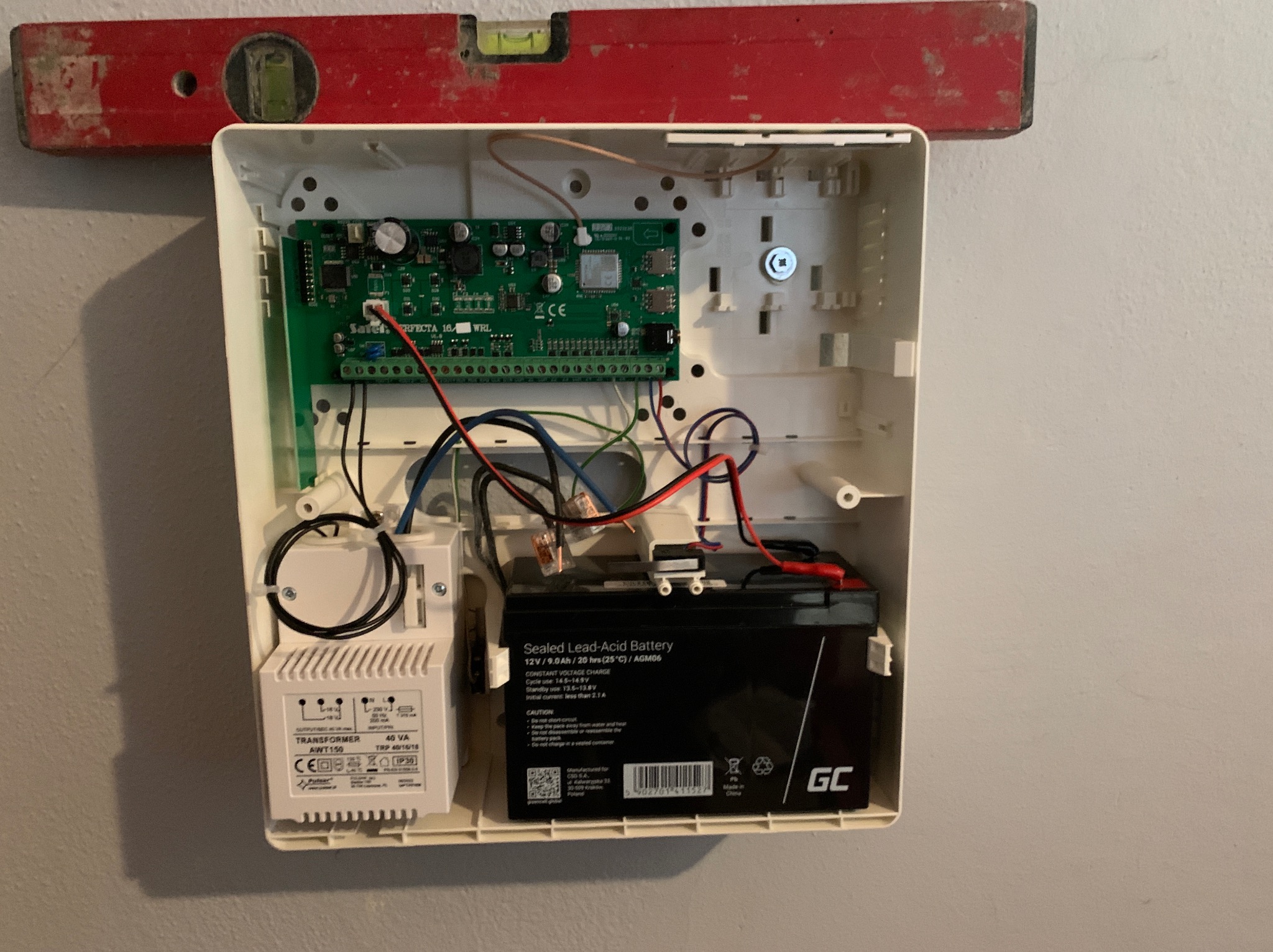 Otwarta obudowa centrali alarmowej z widoczną elektroniką, akumulatorem i transformatorem, poziomica na górnej krawędzi obudowy.