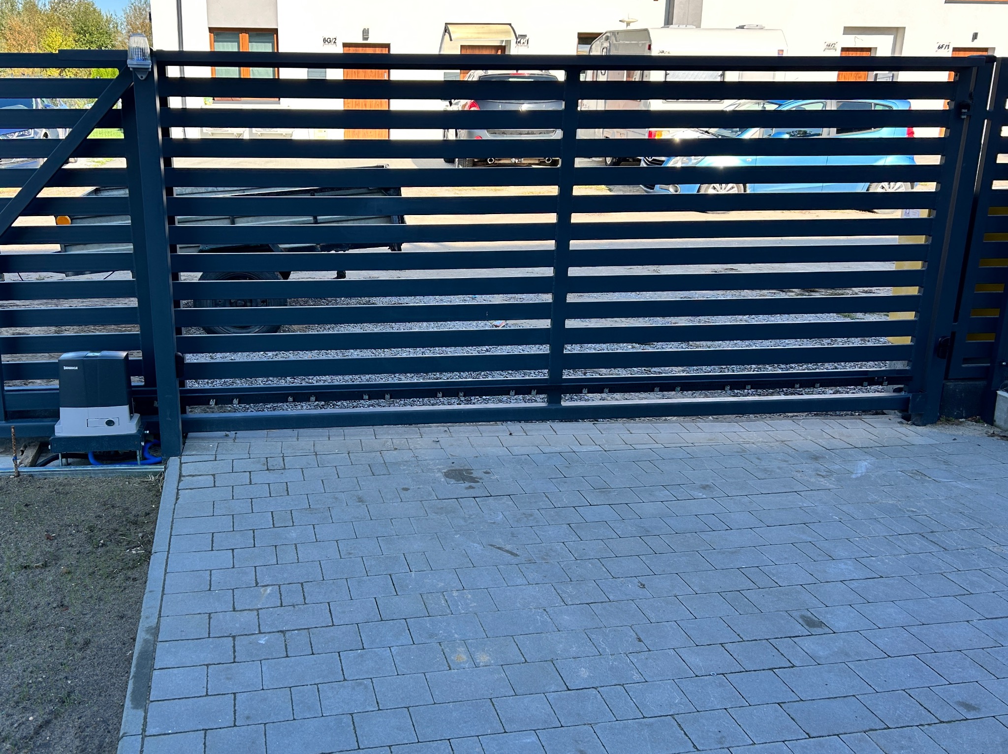 Nowoczesna, pozioma brama wjazdowa z automatyką, szara kostka brukowa na podjeździe, widok na budynek mieszkalny i zaparkowane pojazdy w tle.