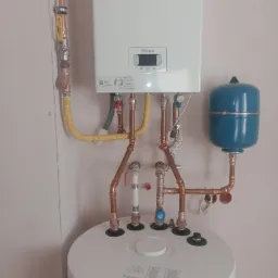 Biały wiszący piec gazowy Beretta z podłączeniami z miedzianych rur, żółtą elastyczną rurą gazową i niebieskim zbiornikiem wyrównawczym, zamontowany nad okrągłym, białym zasobnikiem wody.