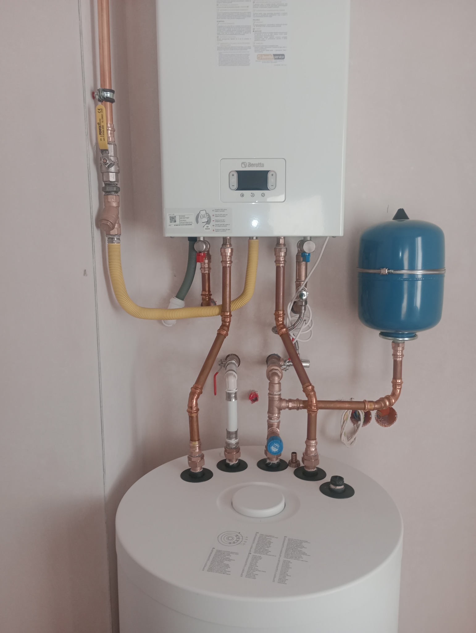 Biały wiszący piec gazowy Beretta z podłączeniami z miedzianych rur, żółtą elastyczną rurą gazową i niebieskim zbiornikiem wyrównawczym, zamontowany nad okrągłym, białym zasobnikiem wody.