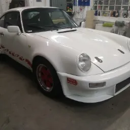 Białe Porsche 911 z czerwonymi akcentami i naklejką '1000 Miglia' na przednim zderzaku, stojące w warsztacie z widocznymi narzędziami i akcesoriami.