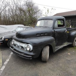Uniloop Sp.zoo - Czarna, matowa, klasyczna ciężarówka pickup z chromowanymi elementami grilla, zaparkowana obok czarnego sportowego samochodu i auta przykrytego plandeką.
