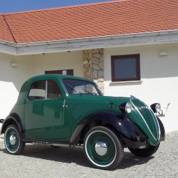 Uniloop Sp.zoo - Odrestaurowany, zielony, zabytkowy samochód z czarnymi błotnikami, zaparkowany przed nowoczesnym, jasnym domem z czerwoną dachówką.